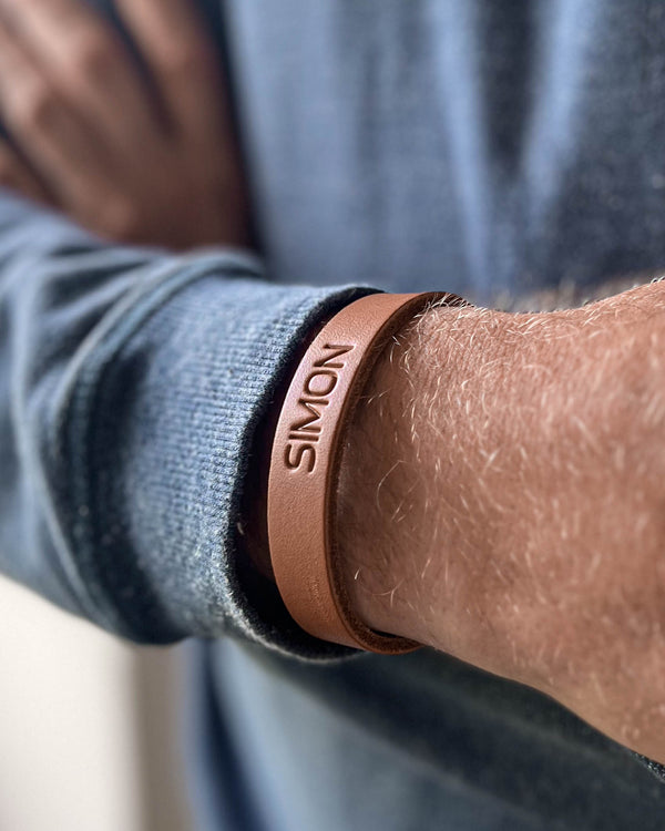 MONO läderarmband i cognac läder med namnet "Simon" graverat, buret runt handleden på man i blå stickad tröja