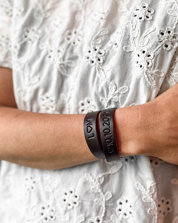 DUO läderarmband i mörkbrunt läder med gravyrerna "L ♡ M" och "♡ 10.10.20 ♡" runt handleden, vit broderad blus