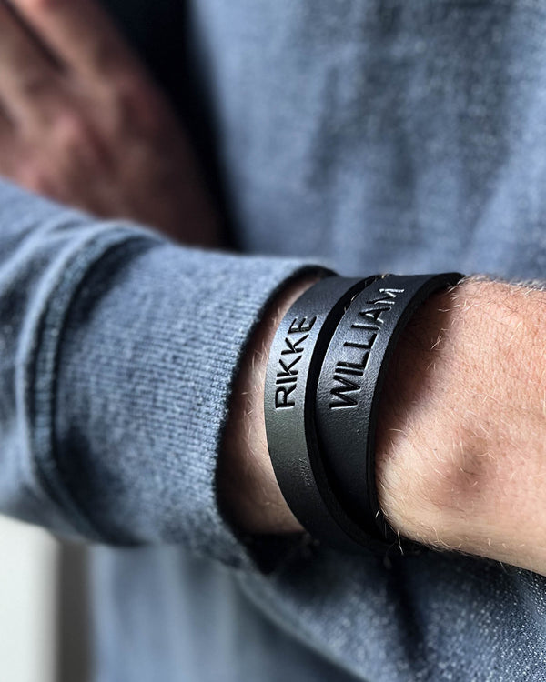 DUO läderarmband i svart läder med namnen "Rikke" och "William" graverade, buret runt manshandled i grå stickad tröja