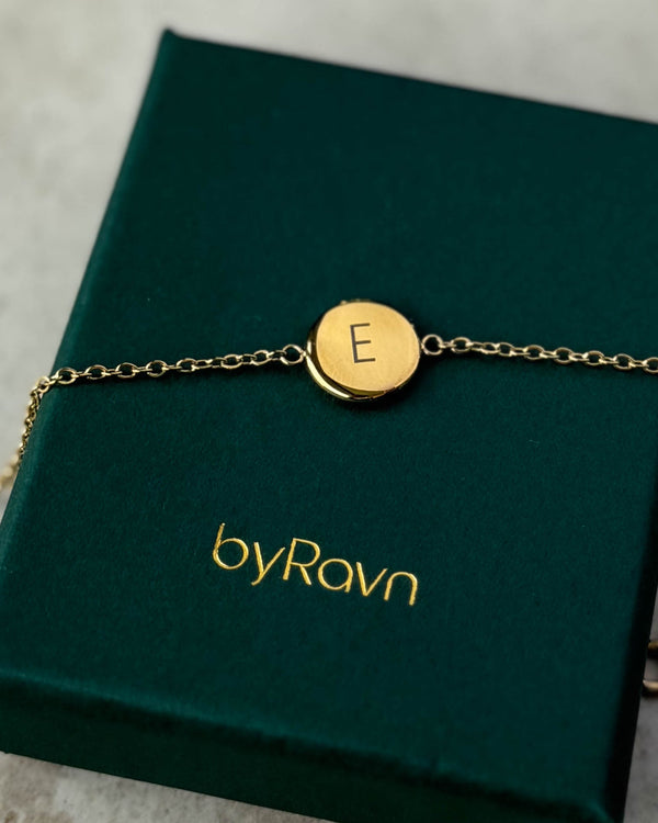 IBI LETTER armband i guldpläterat stål med bokstaven "E" graverad, liggande på mörkgrön byRavn smyckesask
