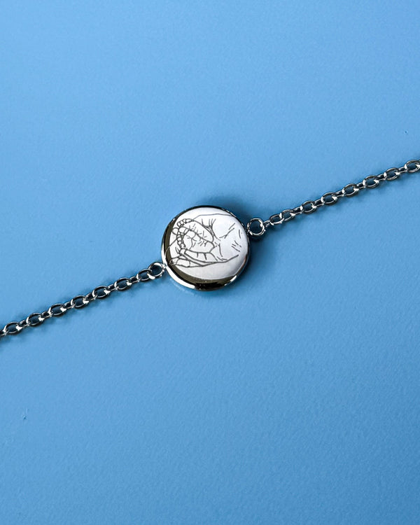 IBI PORTRAIT armband i silverfärgat rostfritt stål med linjeteckning av händer graverad på rund bricka, mot blå bakgrund
