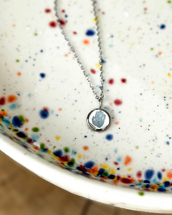 AYA MOTIF halsband i silverfärgat rostfritt stål med ett litet handavtryck graverat på runt hänge, på prickigt keramikfat