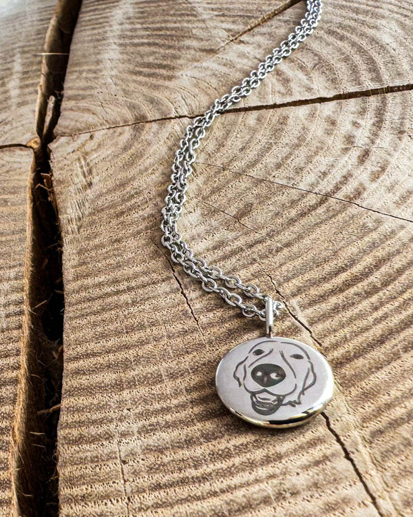 AYA PORTRAIT halsband i silverfärgat rostfritt stål med teckning av en glad hund graverad på litet hänge, på ljust trä med spricka