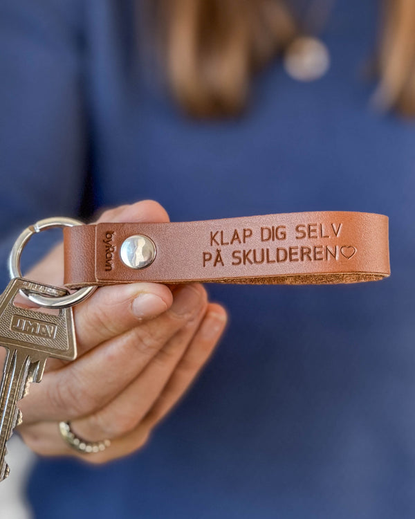 BOND nyckelring i cognac läder med gravyren "Klap dig selv på skulderen" ♡, hålls i handen med nycklar