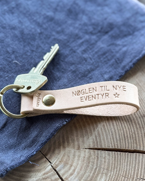 BOND nyckelring i naturfärgat läder med gravyren "Nøglen til nye eventyr" ☆ och mässingsnit, på blått tyg och träbord med nyckel