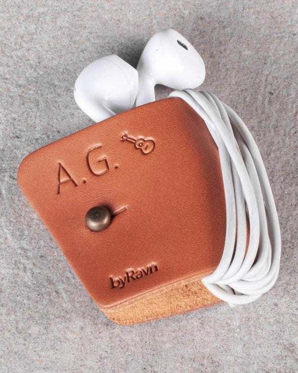 KAX hörlurshållare i cognac läder med initialerna "A.G." och gitarr graverade, med earpods på grå sten
