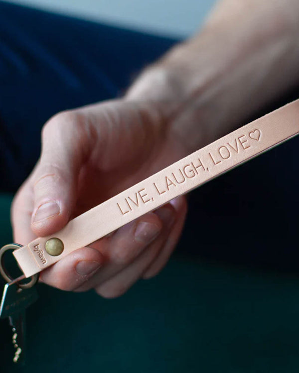LUX nyckelring i naturfärgat läder med mässingsnit och personlig gravyr "Live, laugh, love" ♡, hålls i handen med nyckel