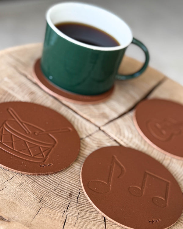 MOKKA glasunderlägg i cognac läder präglade med trumma och noter på träskiva med grön kaffekopp
