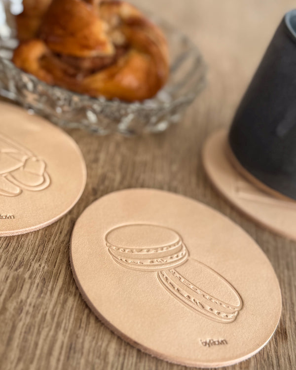 MOKKA kaffe & kaka-glasunderlägg i naturfärgat läder präglat med macarons på träbord vid kanelbulle och svart kopp
