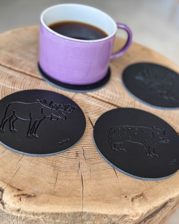 MOKKA jakt-glasunderlägg i svart läder präglade med älg, vildsvin och tjäder på träskiva med lila kaffekopp på underlägg