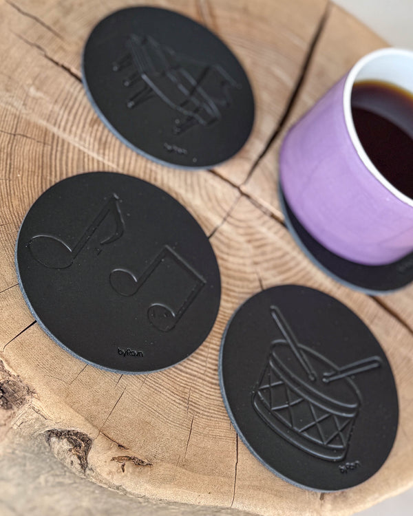 MOKKA glasunderlägg i svart läder präglade med noter, trumma och flygel på träskiva med lila kaffekopp