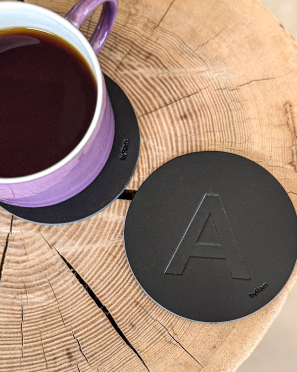 MOKKA glasunderlägg i svart läder präglat med bokstaven "A" på träskiva vid lila kaffekopp på underlägg