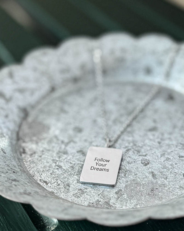 RAZ halsband i silverfärgat rostfritt stål med gravyren "Follow Your Dreams" på rektangulär bricka, på vitpatinerat zinkfat