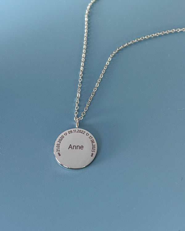 SIA CIRCLE namnhalsband i silverfärgat rostfritt stål med "Anne" och datumen "21.03.2020 ♡ 09.11.2022 ♡ 17.06.2025" i cirkel, på blå botten