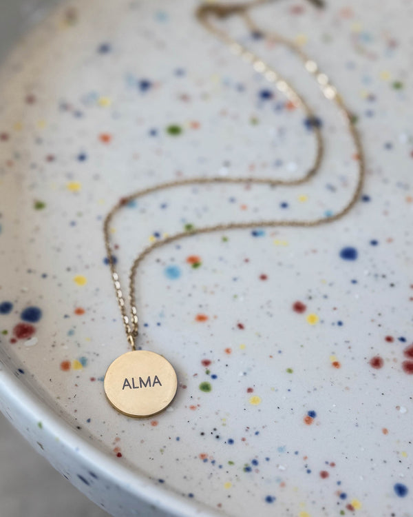 SIA NOTE namnhalsband i guldpläterat stål med namnet "Alma" graverat på runt hänge, på prickigt keramikfat