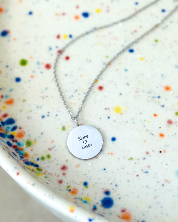 SIA QUOTE namnhalsband i silverfärgat rostfritt stål med gravyren "Signe ♡ Lasse" på runt hänge, på prickigt fat