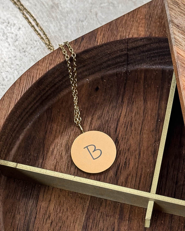 SIA LETTER SCRIPT halsband i guldpläterat stål med bokstaven "B" i handskrift graverad på runt hänge, i träskål med mässingskant