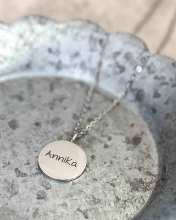 SIA NOTE SCRIPT namnhalsband i silverfärgat rostfritt stål med namnet "Annika" i handskrift graverat, liggande på zinkfat