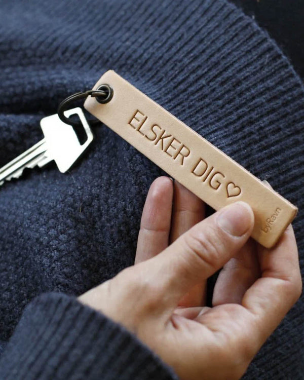 SOLO Medium nyckelring i naturfärgat läder som hålls i handen med nyckel och gravyren "Elsker dig" ♡