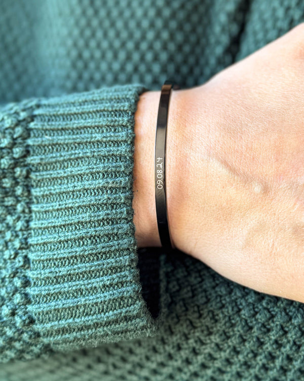 VENUS SCRIPT armband i svart stål med datumet "09.08.24" i handskrift runt handleden, grön stickad tröja
