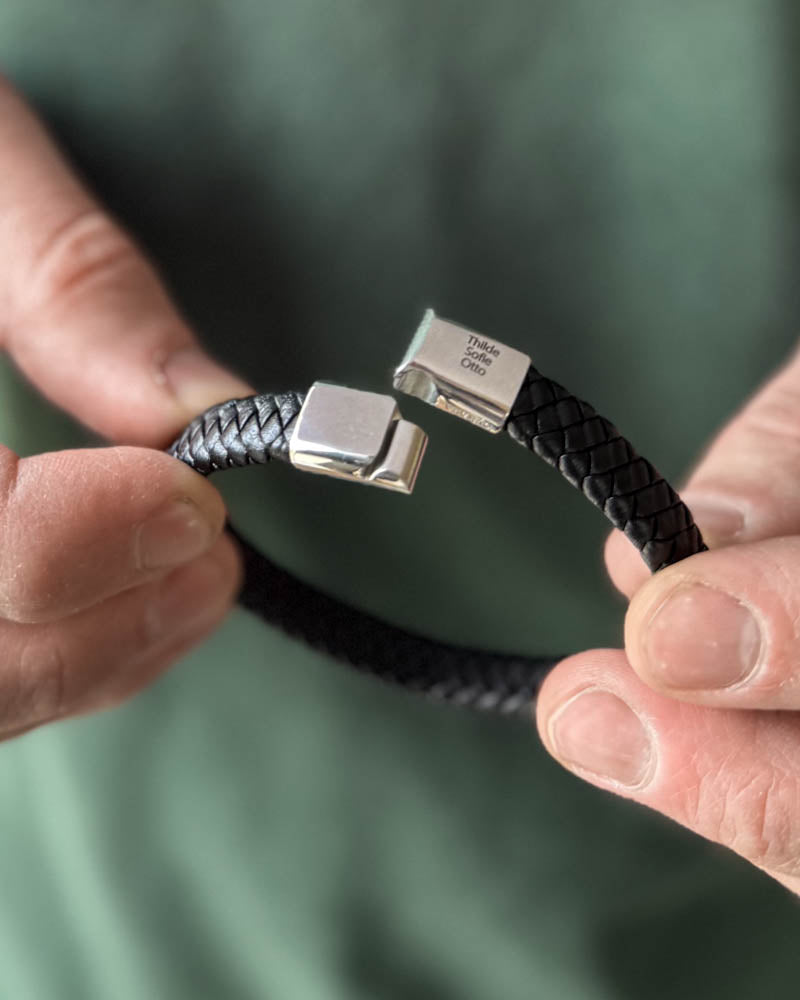 Læderarmbånd – RAY flettet armbånd i sort læder med stållås graveret "Thilde Sofie Otto", holdt i hænderne