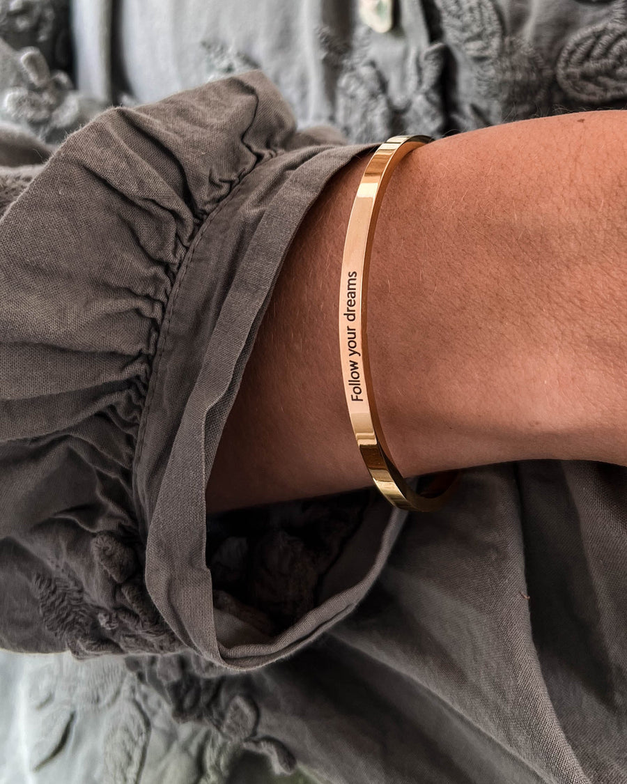 VENUS armband i guldpläterat stål med gravyren "Follow your dreams" buret runt handleden