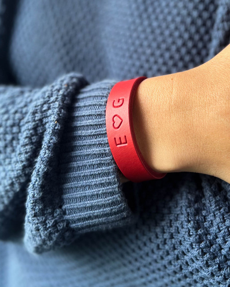 MONO läderarmband i rött läder med initialerna "E ♡ G" graverade runt handleden, buret till blå stickad tröja