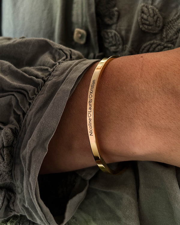 VENUS armband i guldpläterat stål med gravyren "Nicoline ♡ Laurits ♡ Kristian" runt handleden, grå blus