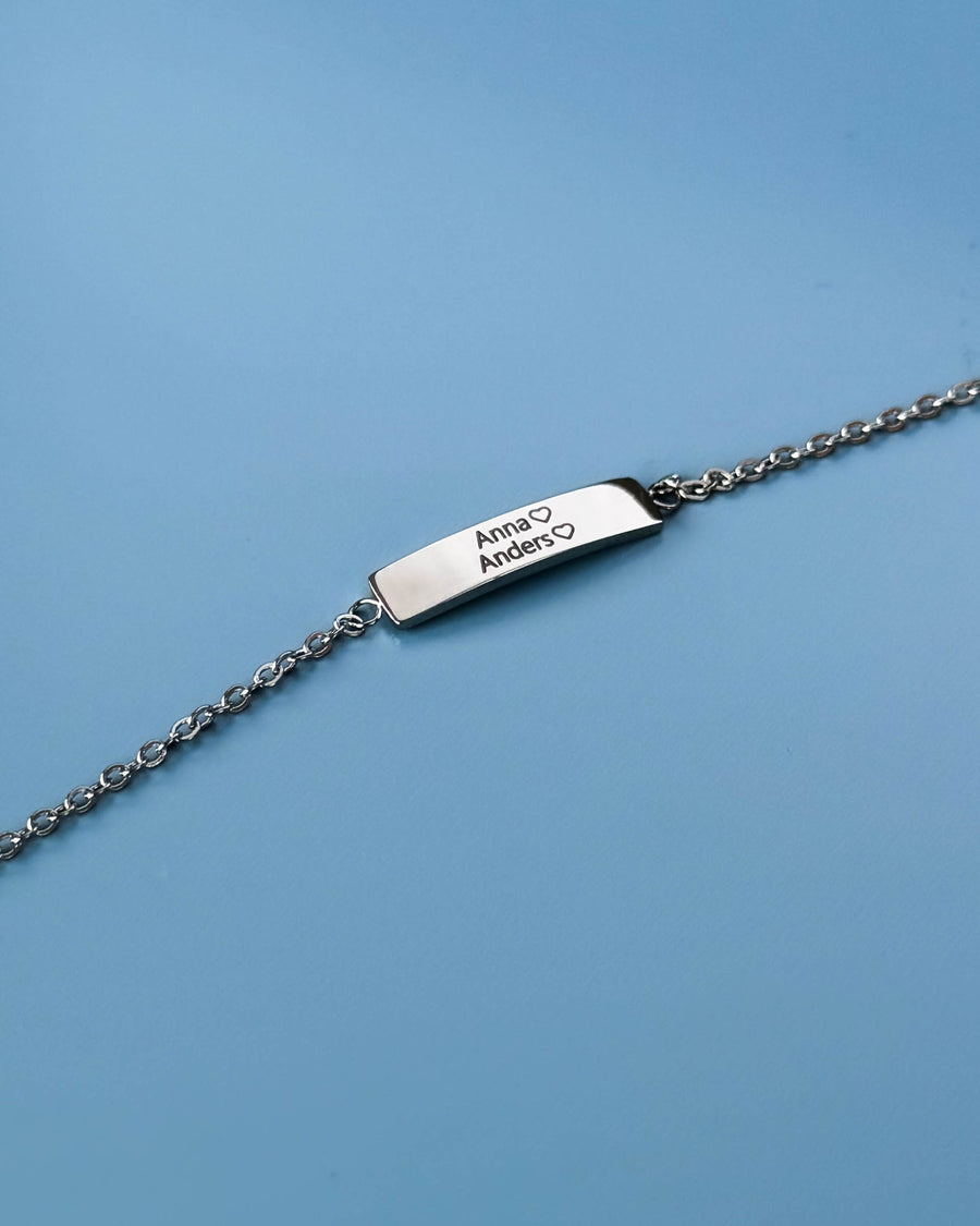 EVI namnarmband i silverfärgat rostfritt stål med namnen "Anna" ♡ "Anders" ♡ graverade, mot ljusblå bakgrund