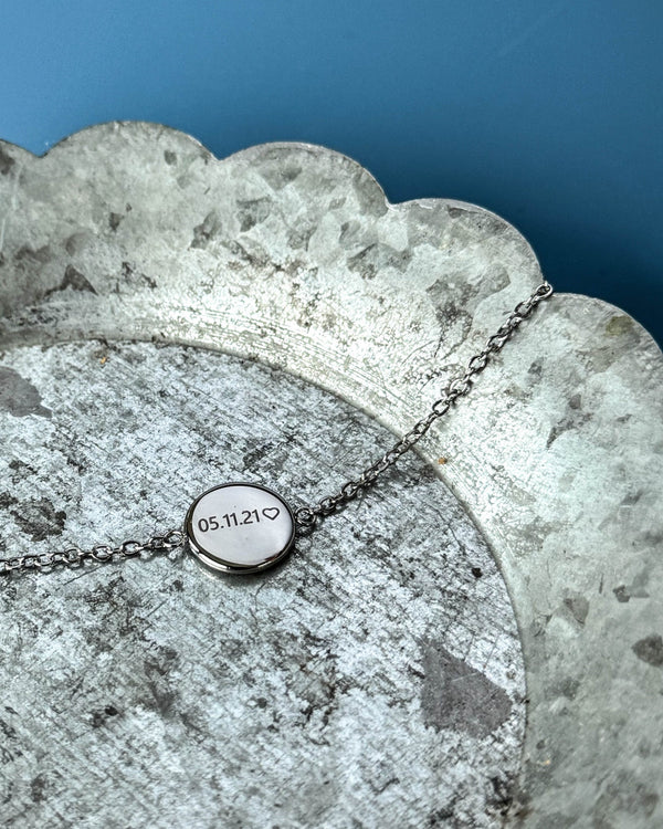 IBI NOTE armband i silverfärgat rostfritt stål med "05.11.21" ♡ graverat på rund bricka, på zinkfat mot blå bakgrund