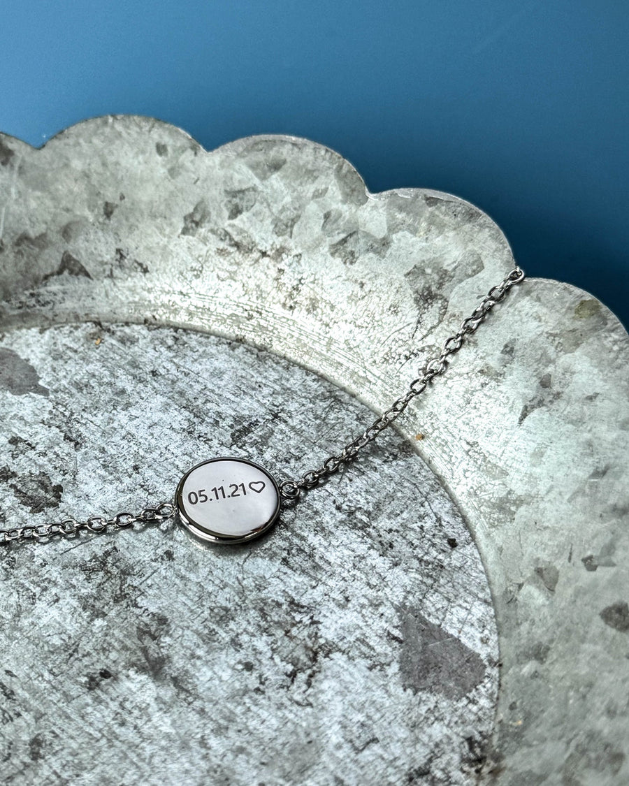 IBI NOTE armband i silverfärgat rostfritt stål med "05.11.21" ♡ graverat på rund bricka, på zinkfat mot blå bakgrund