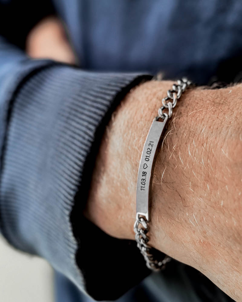 ARA armband i silverfärgat rostfritt stål med datumen "11.03.18 ♡ 01.02.21" graverade, runt en mans handled, blå sweatshirt
