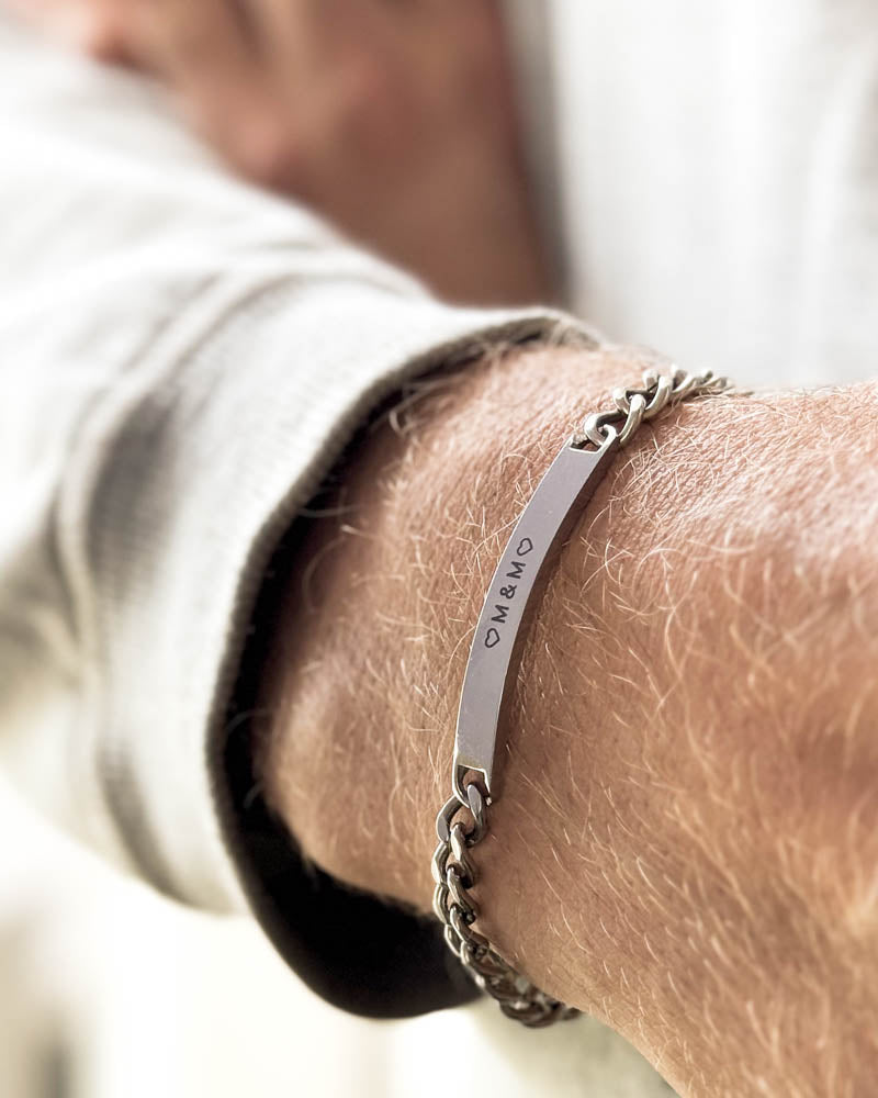 ARA armband i silverfärgat rostfritt stål med initialerna "♡ M & M ♡" graverade runt handleden, vit sweatshirt