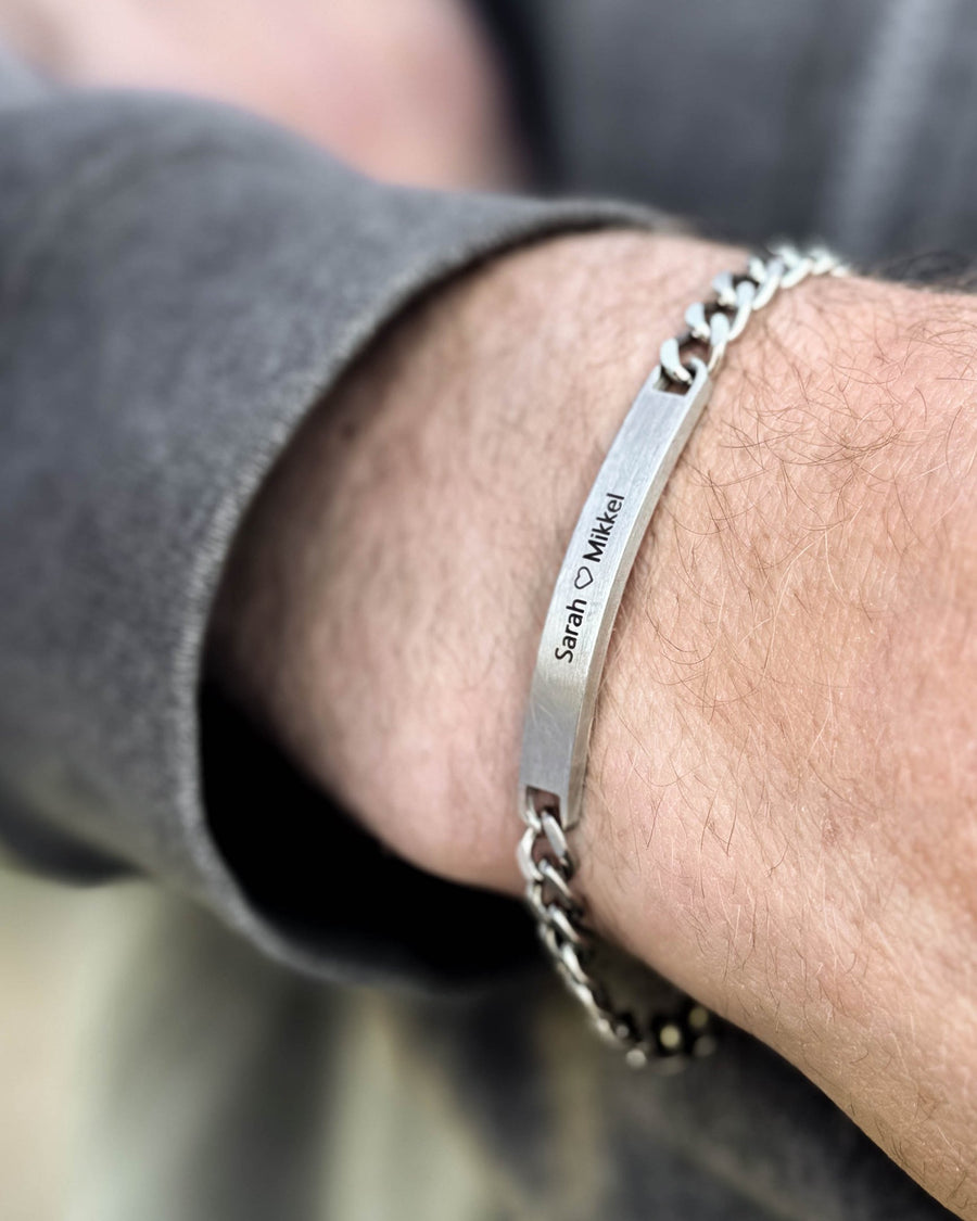 ARA armband i borstat silverfärgat rostfritt stål med gravyren "Sarah ♡ Mikkel" buret runt mannens handled