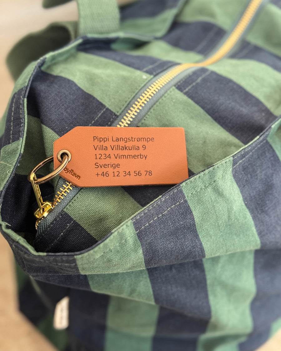 ATLAS bagagebricka i läder, här i cognac, med graverad adress "Pippi Langstrømpe, Villa Villakulla 9" på grönblå randig väska