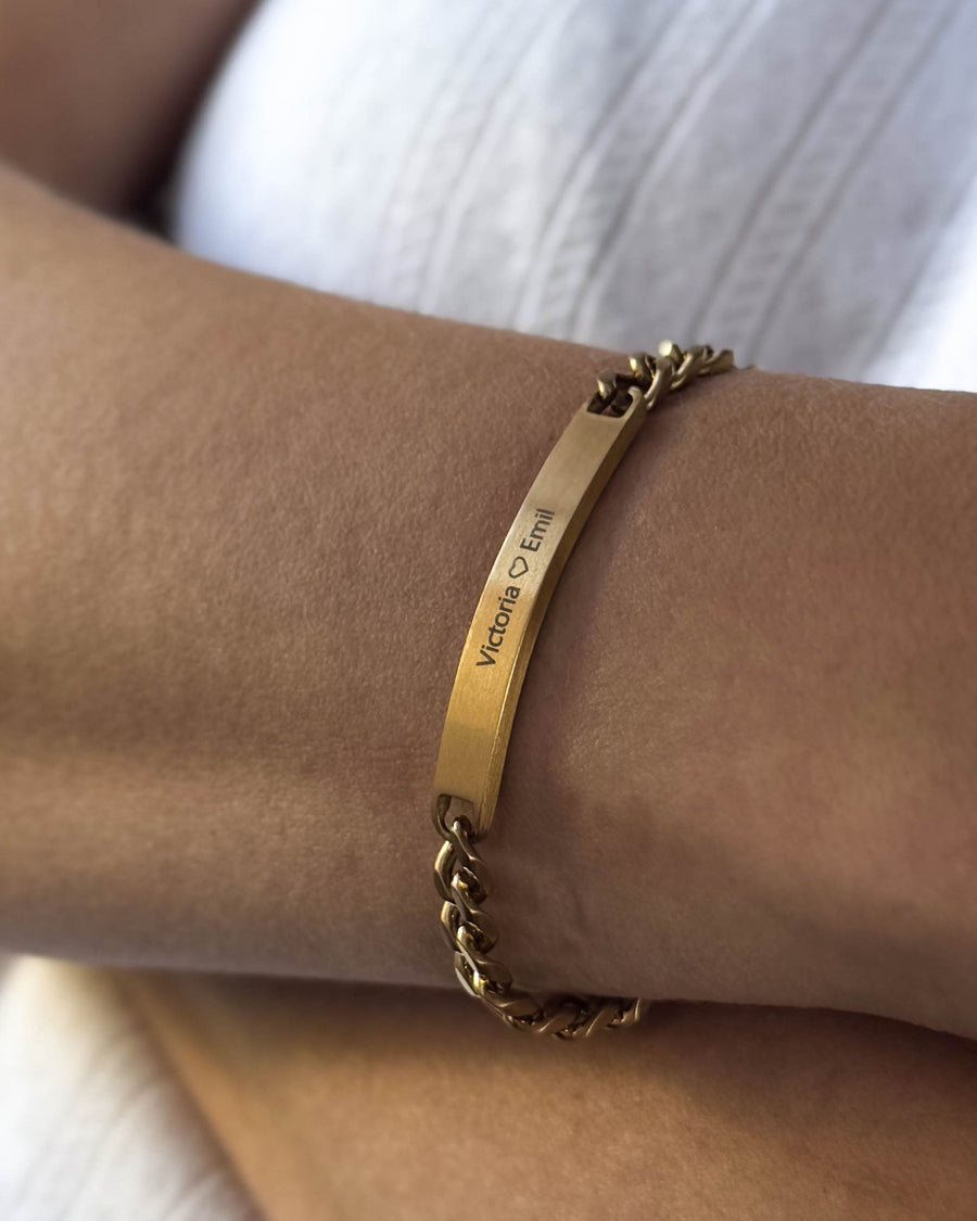AVA armband i borstat guldpläterat stål med personlig gravyr "Victoria ♡ Emil" buret runt handleden
