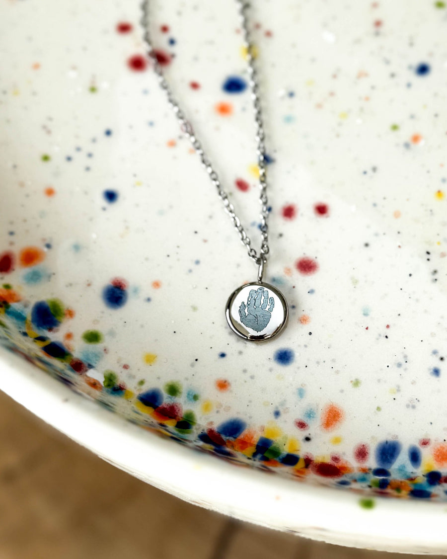 AYA MOTIF halsband i silverfärgat rostfritt stål med ett litet handavtryck graverat på runt hänge, på prickigt keramikfat