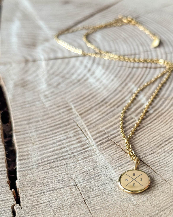 AYA X halsband i guldpläterat stål med initialerna "B N S E" graverade i kors på litet runt hänge, på ljust trä med spricka