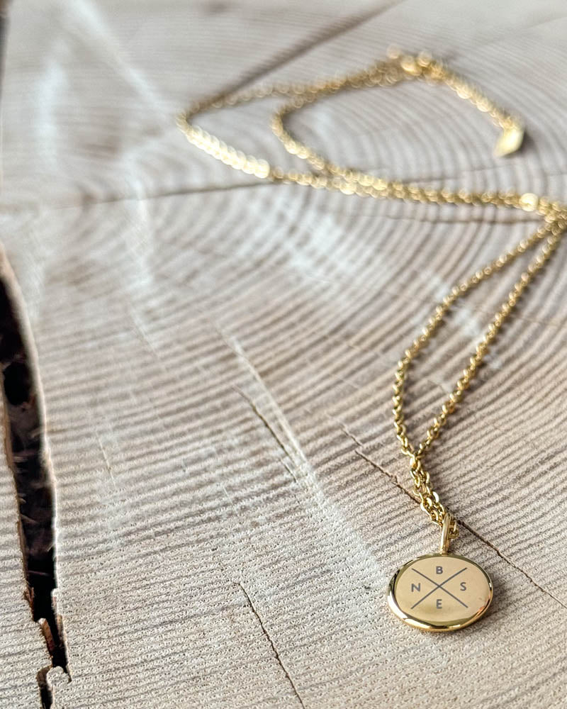 AYA X halsband i guldpläterat stål med initialerna "B N S E" graverade i kors på litet runt hänge, på ljust trä med spricka