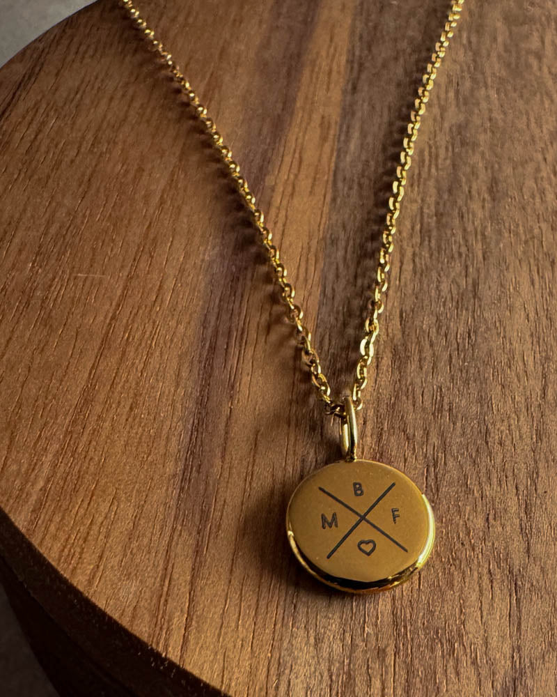 AYA X halsband i guldpläterat stål med bokstäverna "B M F ♡" graverade i kors på litet hänge, på mörk träskiva