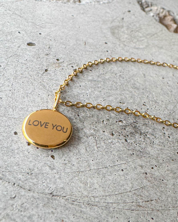 AYA NOTE halsband i guldpläterat stål med gravyren "Love you" på litet runt hänge, liggande på betong
