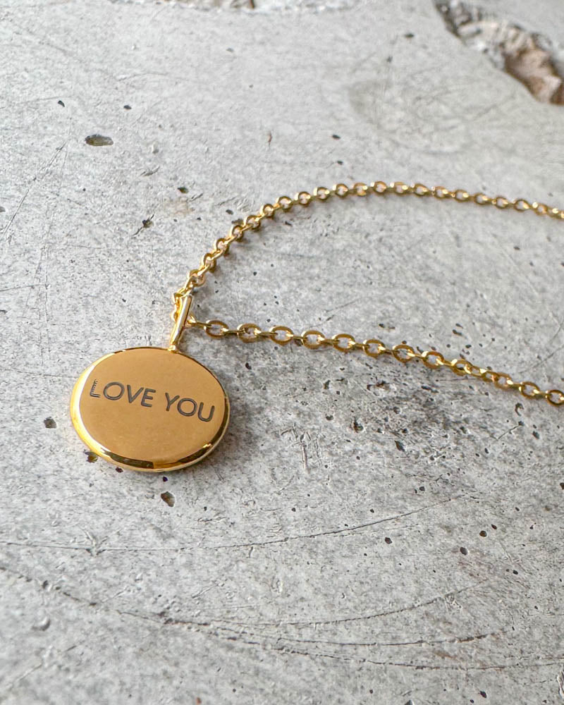 AYA NOTE halsband i guldpläterat stål med gravyren "Love you" på litet runt hänge, liggande på betong