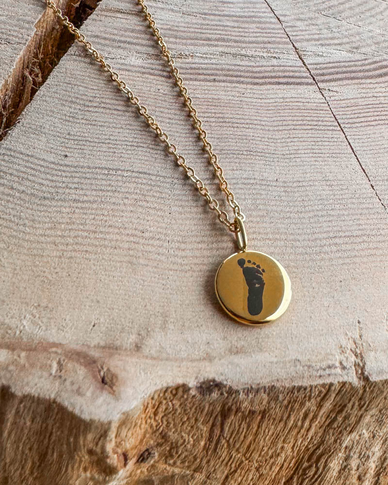AYA MOTIF halsband i guldpläterat stål med babyns fotavtryck graverat på litet hänge, på ljust trä med spricka