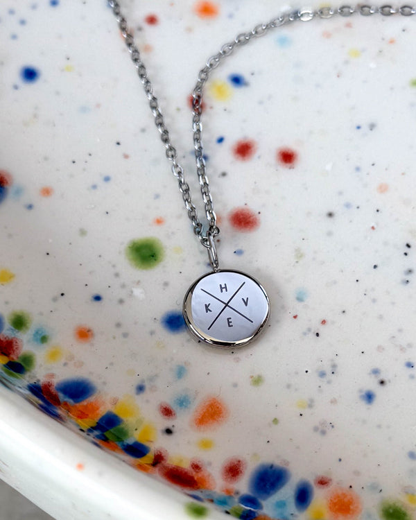 AYA X halsband i silverfärgat rostfritt stål med initialerna "H K V E" graverade i kors på litet hänge, på prickigt keramikfat
