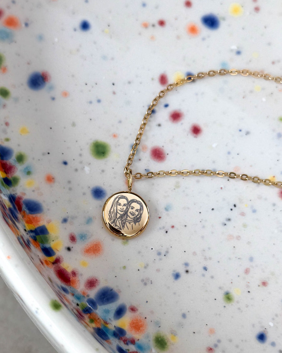 AYA PORTRAIT halsband i guldpläterat stål med linjetecknat porträtt av två kvinnor på litet hänge, på prickigt keramikfat