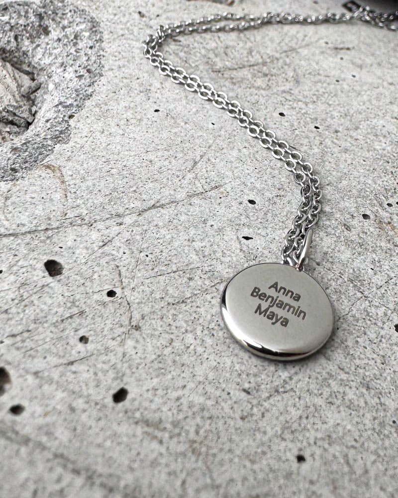 AYA QUOTE namnhalsband i silverfärgat rostfritt stål med namnen "Anna Benjamin Maya" graverade på litet runt hänge, på betong