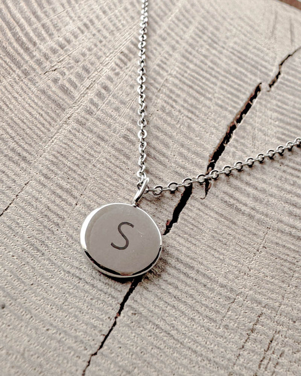 AYA LETTER halsband i silverfärgat rostfritt stål med bokstaven "S" graverad på litet runt hänge, på ljust trä med spricka