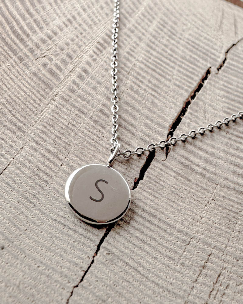 AYA LETTER halsband i silverfärgat rostfritt stål med bokstaven "S" graverad på litet runt hänge, på ljust trä med spricka