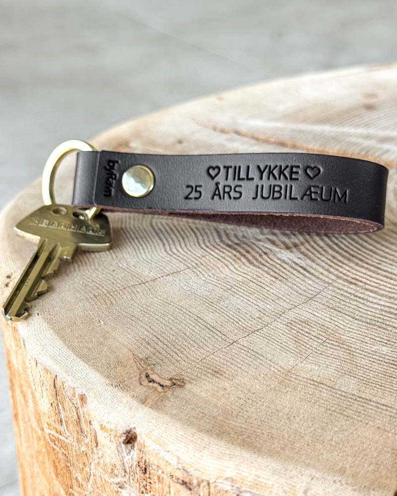 BOND nyckelring i mörkbrunt läder med gravyren "Tillykke 25 års jubilæum" ♡ och mässingsnit, liggande på trästubbe med nyckel