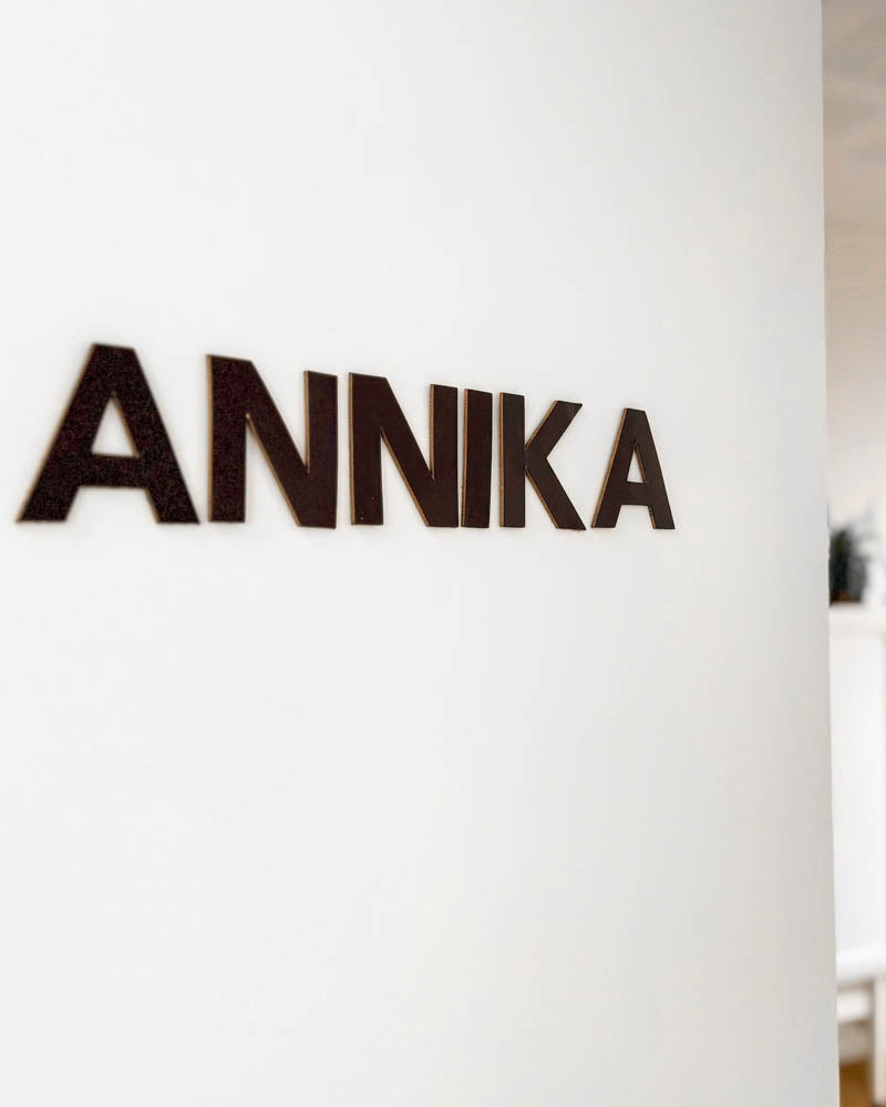BOX bokstäver i mörkbrunt läder med namnet "Annika" monterade på vit vägg